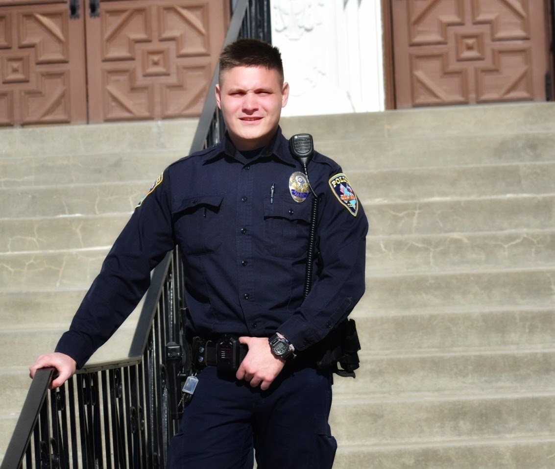 A police officer in a dark uniform stands outside on concrete steps, holding a black railing with one hand and looking at the camera. The background shows wooden doors with geometric patterns.