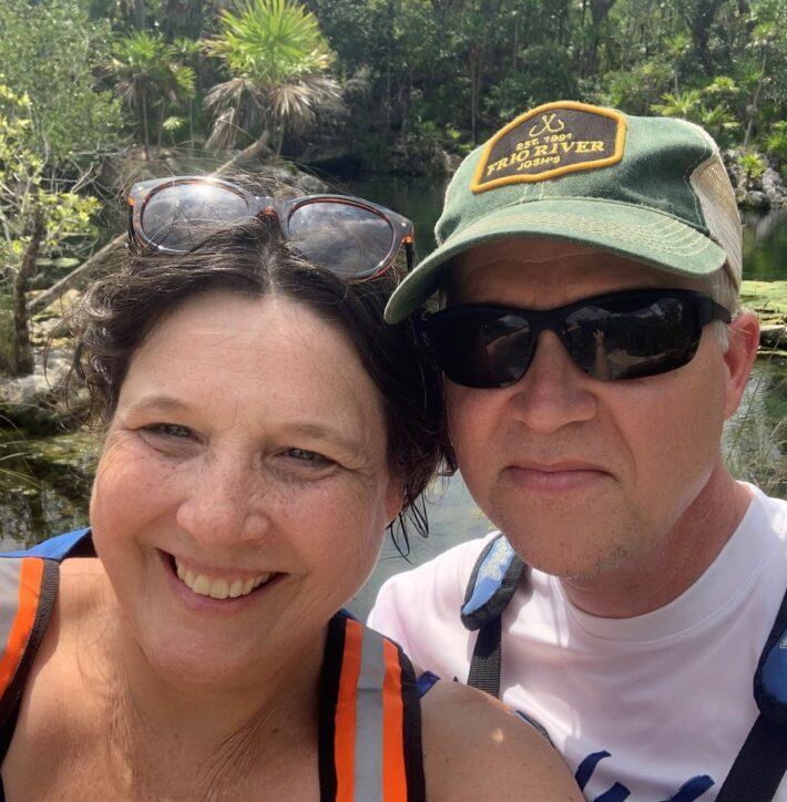 A smiling woman and a man wearing sunglasses and a green cap take a selfie outdoors, with lush greenery and water in the background. The woman has sunglasses resting on her head.