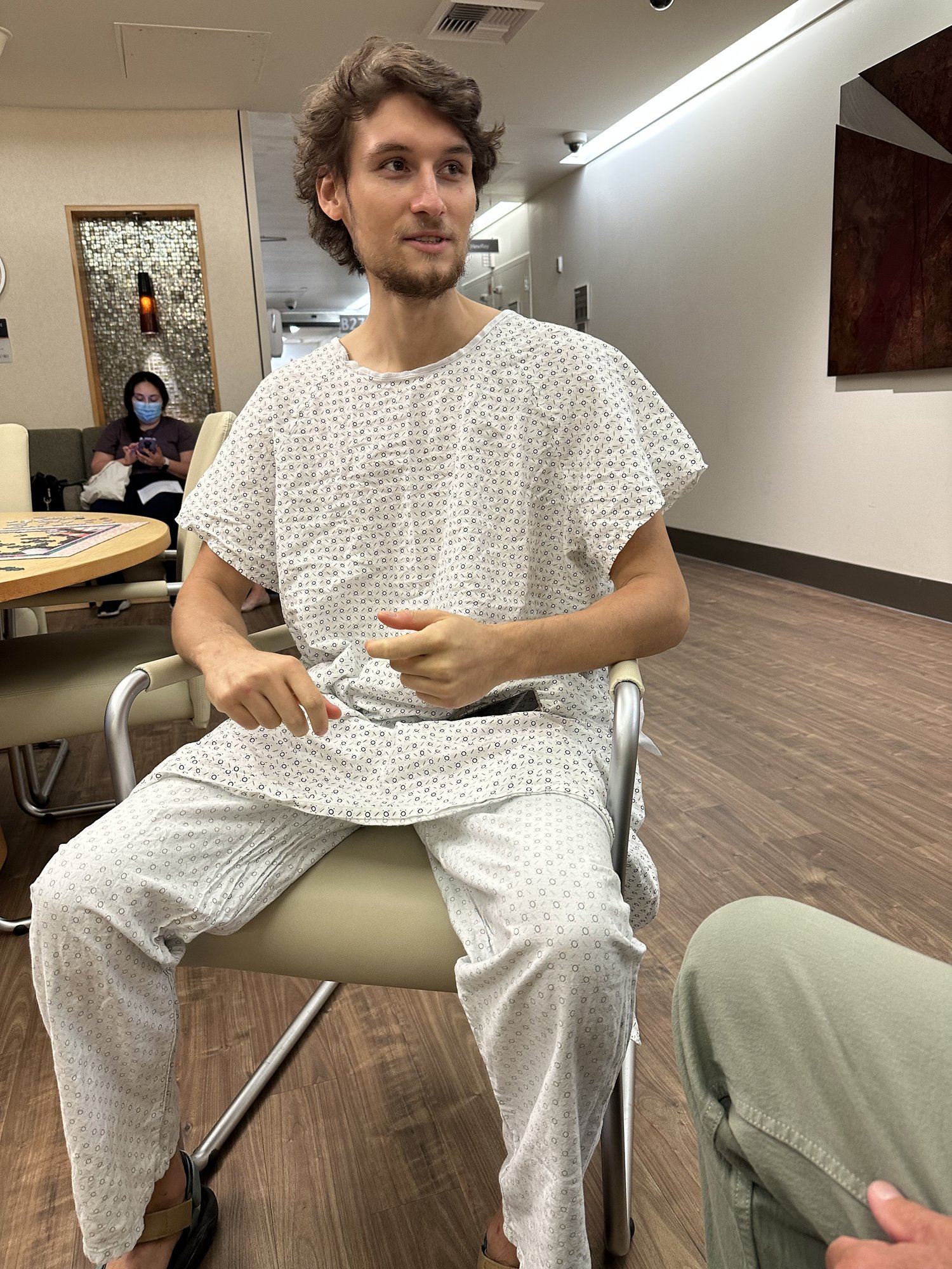 A young man wearing a hospital gown sits on a chair in a waiting room with wood floors. He appears to be talking. Another person and a table with magazines are visible in the background.
