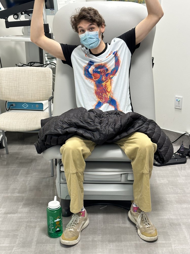 A young person wearing a mask and a graphic monkey t-shirt sits on a medical exam chair with arms raised. A black jacket is on their lap, and a green water bottle is on the floor next to their feet.