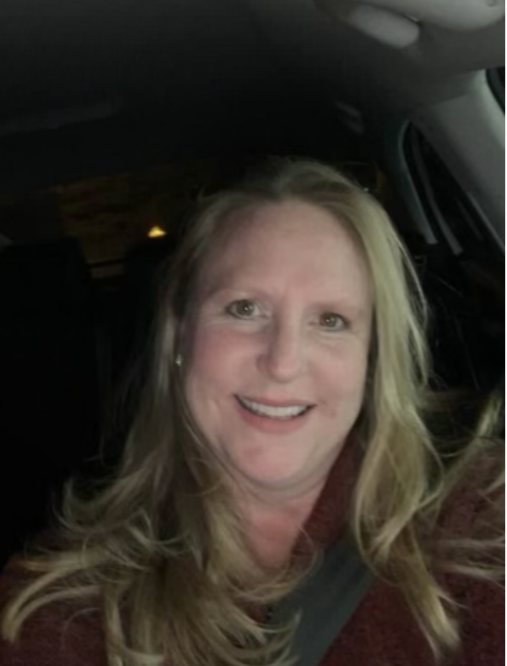 A woman with long blonde hair smiles at the camera while sitting in the driver's seat of a car at night. She is wearing a dark top and a seatbelt. The car interior is dimly lit.