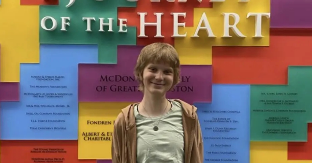 A smiling person with short hair stands in front of a colorful wall with raised text that reads "JOURNEY OF THE HEART" and various names and foundations displayed on colored panels.