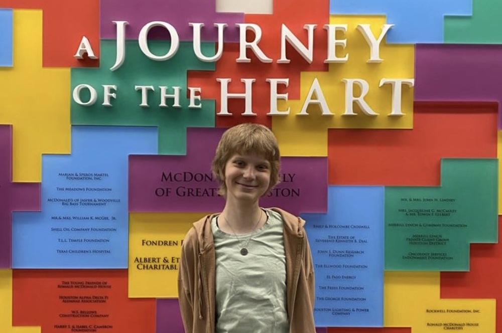A person with short light brown hair smiles while standing in front of a colorful wall with blocks and the words “A Journey of the Heart” displayed in large white letters.
