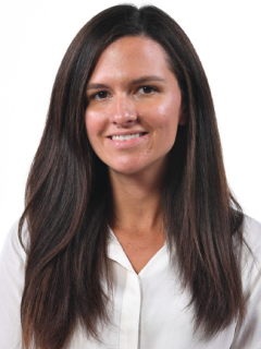 A woman with long straight brown hair, wearing a white blouse, smiles softly at the camera against a plain white background.