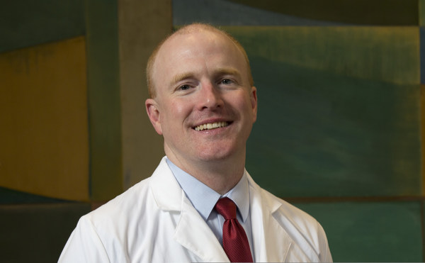 A person with short, light hair is smiling, wearing a white lab coat, a blue shirt, and a red tie. The background features abstract geometric patterns in shades of green and yellow.