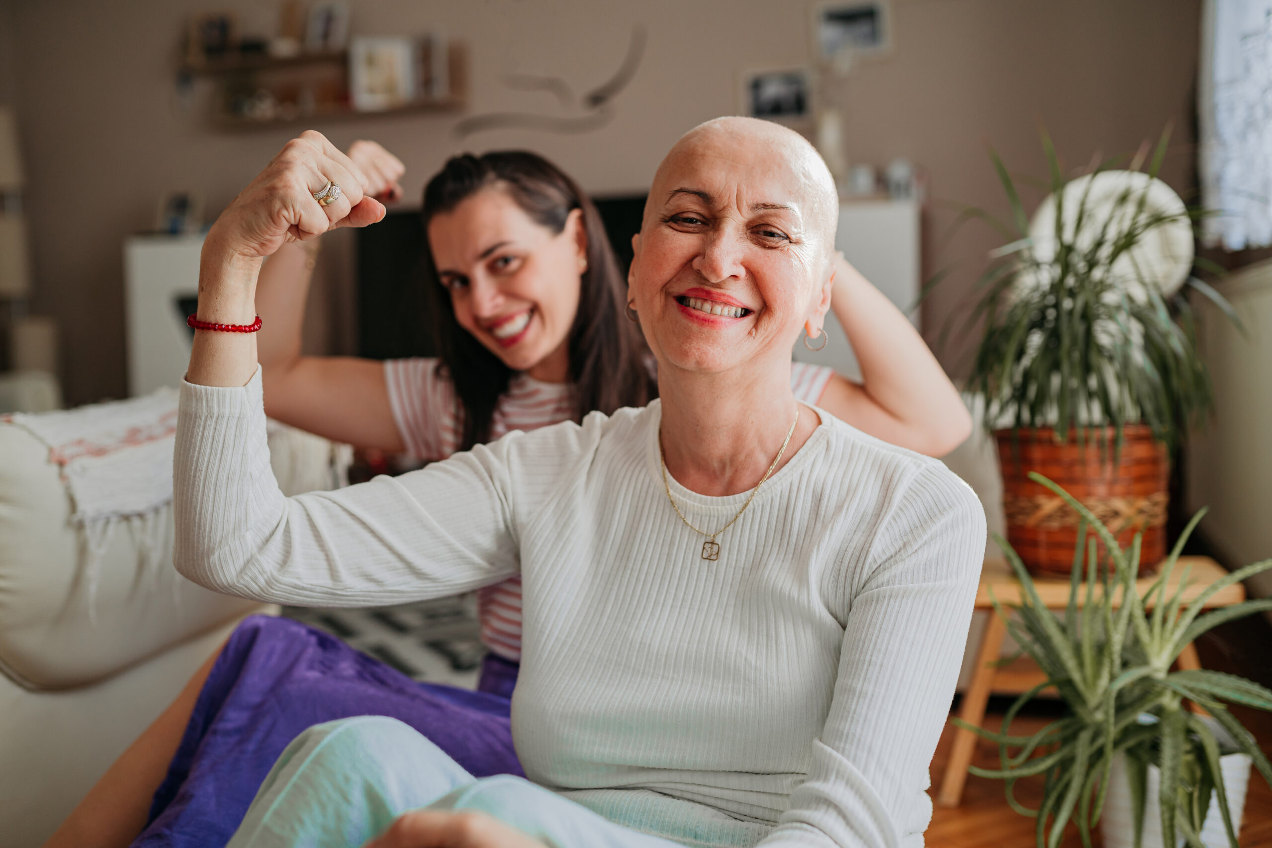 A bald woman and a younger woman sit together on a couch, both smiling and flexing their arms in a gesture of strength. The atmosphere is warm and supportive, with indoor plants in the background.