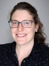A woman with curly brown hair, wearing glasses and a black shirt with white dots, smiles at the camera against a plain gray background.