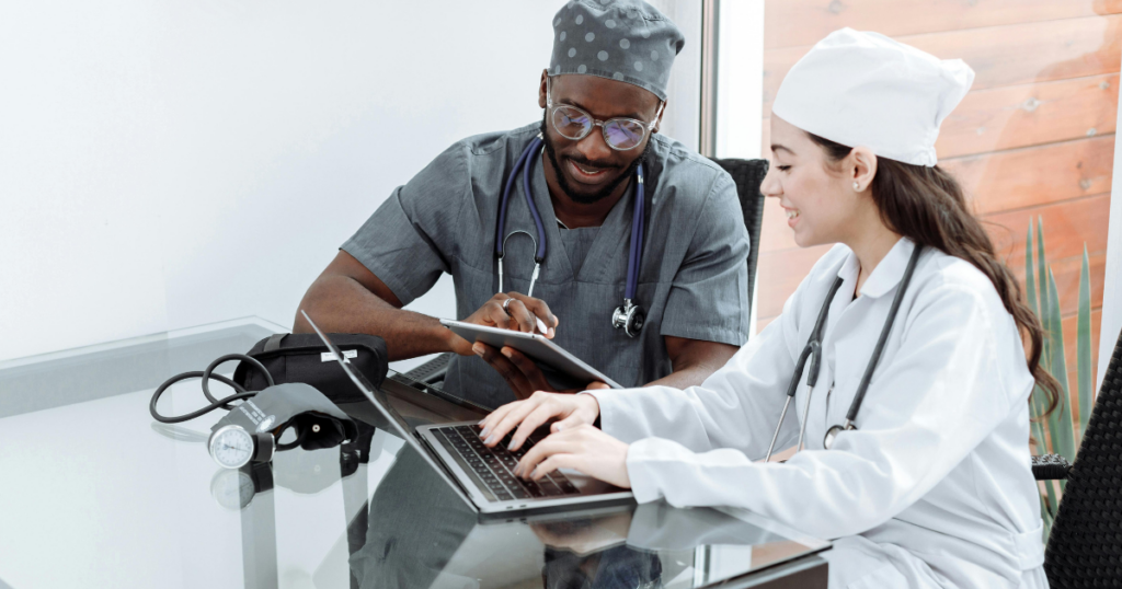 Two healthcare professionals, one in grey scrubs and the other in a white lab coat, sit at a glass desk. They are discussing something on a tablet and laptop, with a blood pressure monitor nearby.