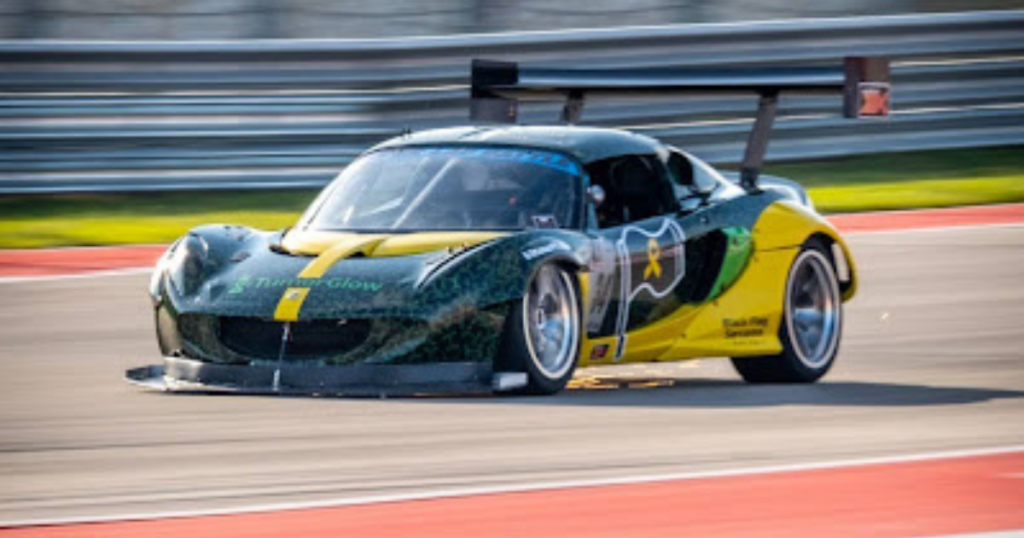 A green and yellow race car speeds around a track, with motion blur showing its fast movement. Barriers and a red-and-white curb are visible in the background.