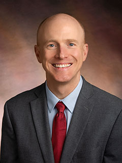 A smiling bald man in a gray suit, light blue shirt, and red tie poses for a professional portrait against a softly blurred brown background.