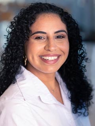 A woman with long curly black hair smiles warmly at the camera. She is wearing a white collared shirt and gold hoop earrings, with a blurred background behind her.