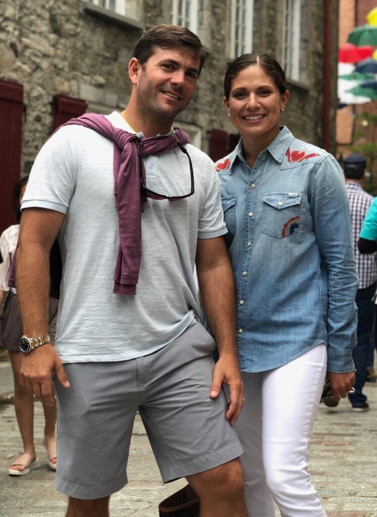 A man in a light shirt and shorts stands next to a woman in a denim shirt and white pants. They are smiling outdoors on a cobblestone street with stone buildings and other people in the background.