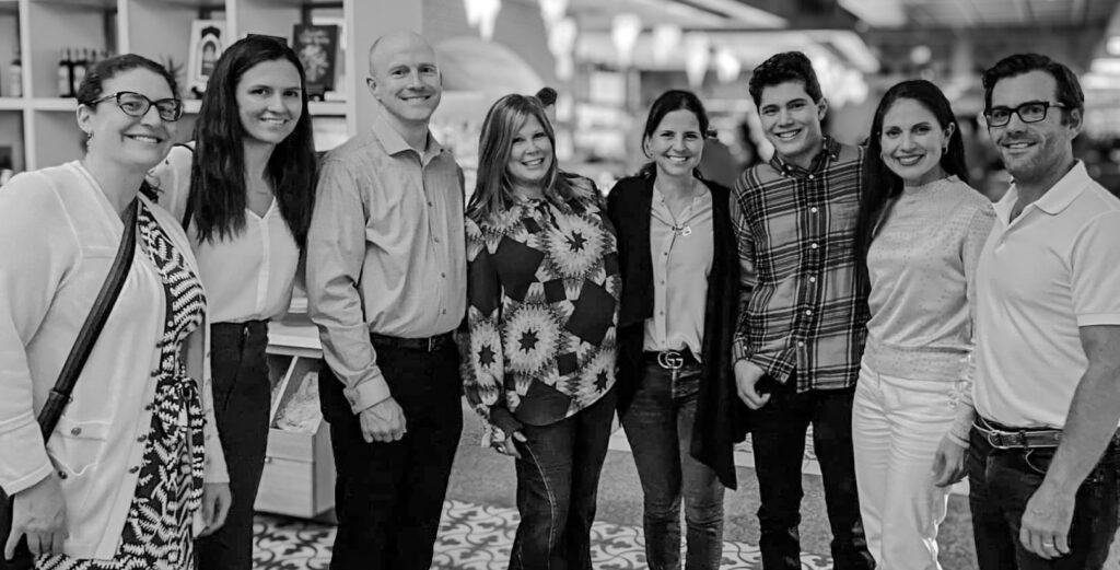 A black and white group photo of eight people standing closely together, smiling. They are indoors, with shelves and a patterned floor visible. The group includes both men and women, dressed in casual and semi-casual attire.