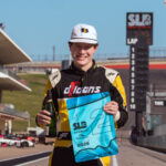 A young race car driver in a black and yellow jumpsuit stands on a racetrack holding a bottle and a blue award flag, smiling. Race cars, a grandstand, and a large American flag are visible in the background.