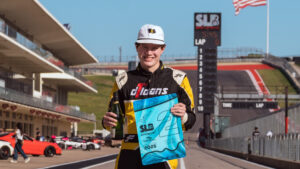 A young race car driver in a black and yellow jumpsuit stands on a racetrack holding a bottle and a blue award flag, smiling. Race cars, a grandstand, and a large American flag are visible in the background.