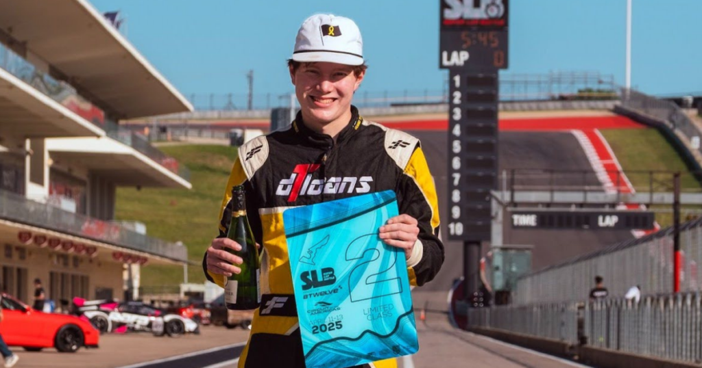 A smiling race car driver stands on a racetrack at Super Lap Battle 2025, holding a bottle and a blue award banner with "2025" and a "2" on it. FASTx Autosport cars and a scoreboard are visible in the background under a clear sky.