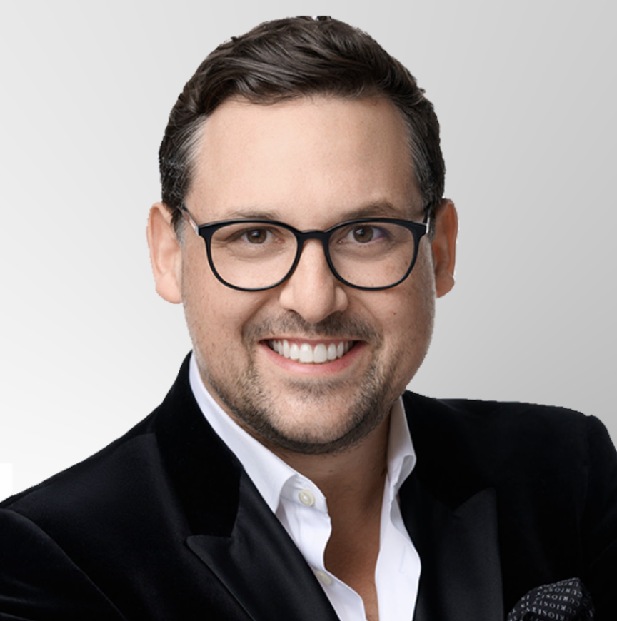 A smiling man with short dark hair, wearing black glasses, a black velvet jacket, and a white shirt, posing against a light grey background.