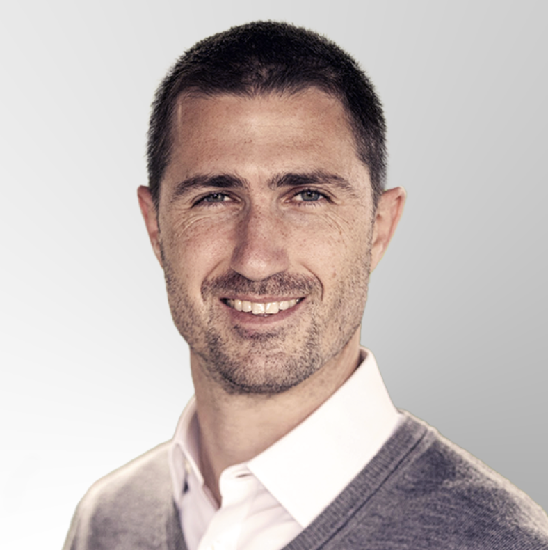 Smiling man with short brown hair and light stubble, wearing a light pink collared shirt and gray sweater, posing against a light gray gradient background.