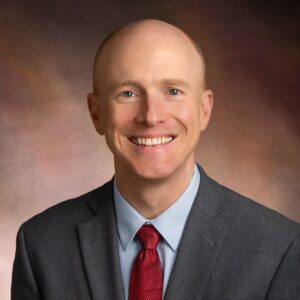 A smiling man in a gray suit, light blue shirt, and red tie poses for a professional headshot against a brown, softly blurred background.