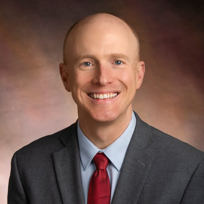 A smiling man in a gray suit, light blue shirt, and red tie poses for a professional headshot against a brown, softly blurred background.