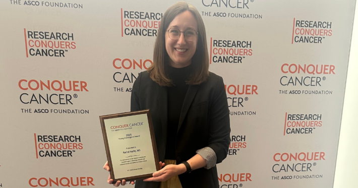 A smiling woman with glasses holds an award plaque in front of a backdrop featuring “CONQUER CANCER,” “RESEARCH CONQUERS CANCER,” and Synovial Sarcoma Foundation logos from The ASCO Foundation.