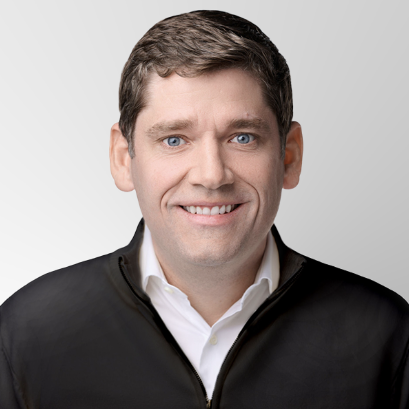 A man with short brown hair and blue eyes, wearing a white collared shirt and a black zip-up jacket, smiling in front of a light gray background.