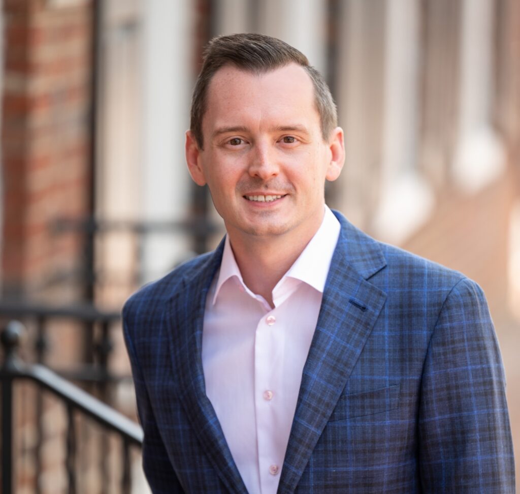 A man in a blue plaid suit jacket and light pink shirt stands outside, smiling, with a blurred background of brick buildings and railing.