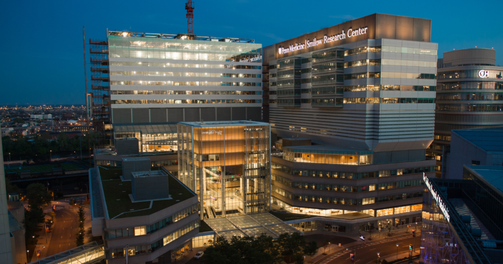 A modern, multi-story medical research center is illuminated at dusk, with large glass windows and well-lit interiors. The building is labeled “Houston Methodist Research Institute.” City lights are visible in the background.