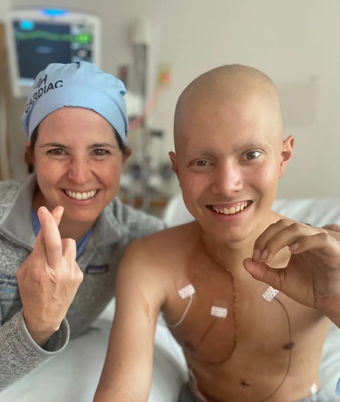 A smiling medical professional wearing a blue cap and a young patient with a bald head and medical electrodes on their chest pose together in a hospital room, both making encouraging hand gestures.