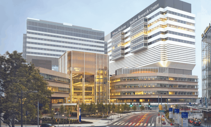 A modern, multi-story medical and research center with glass and metal facades, brightly lit interiors, and surrounding trees. A street with crosswalks and traffic signs runs in front of the building.