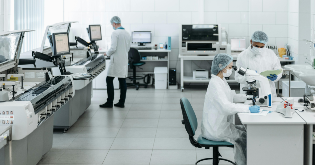 Three scientists in white lab coats and hairnets work in a modern laboratory; one looks into a microscope, another reviews documents, and a third operates equipment in the background.