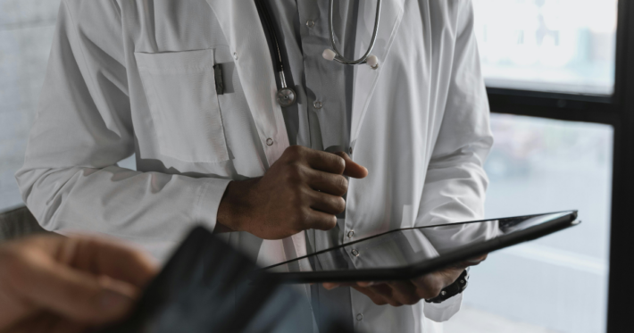 A doctor in a white coat with a stethoscope around their neck holds a digital tablet, standing near a window. Only the upper body is visible. Another hand is blurred in the foreground.