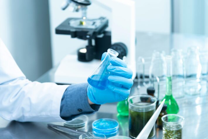 A scientist wearing blue gloves holds a flask with blue liquid in a laboratory, surrounded by various glassware and a microscope in the background.