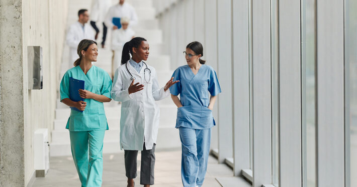 Three medical professionals walk and talk in a bright hallway. One wears a white coat with a stethoscope, while the others wear scrubs. Two more people in white coats are visible on the stairs in the background.