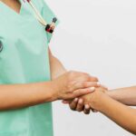 A nurse in green scrubs with a stethoscope gently holding the hands of a patient, offering comfort and support against a plain white background.