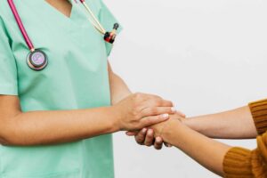 A nurse in green scrubs with a stethoscope gently holding the hands of a patient, offering comfort and support against a plain white background.