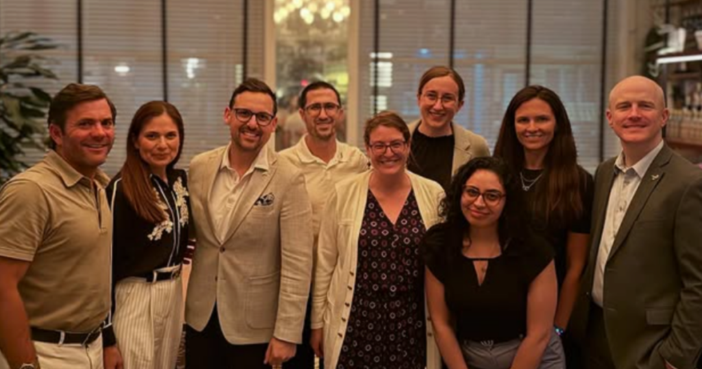 A group of nine adults, both men and women, stand closely together indoors at a Synovial Sarcoma event, smiling at the camera. They are dressed in business or business casual attire, with a warmly lit background featuring blinds and plants.