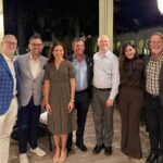 Seven people, four men and three women, stand together smiling on a covered outdoor patio at night. Dressed in business or smart casual attire, they appear to be attending a Connective Tissue Oncology Society (CTOS) event on synovial sarcoma.