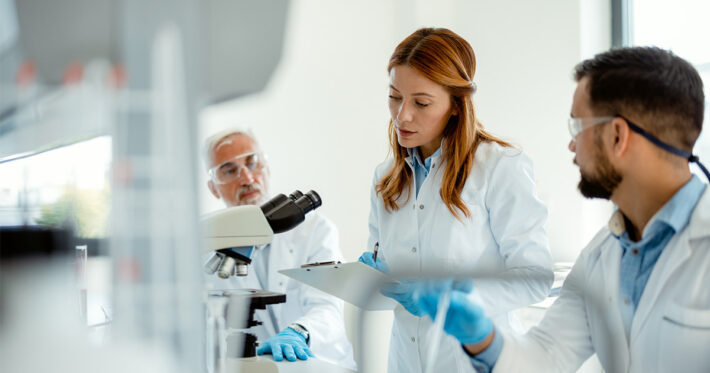 Three scientists in lab coats and safety glasses work together in a laboratory. One woman holds a clipboard and takes notes while the two men, one beside a microscope, listen and discuss results.