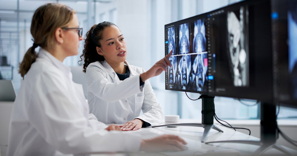 Two doctors in white lab coats examine and discuss medical scan images displayed on computer monitors in a modern, bright medical office or laboratory setting.