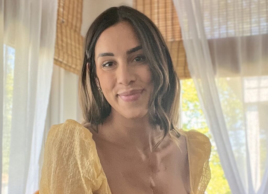 A woman with shoulder-length brown hair and a yellow top smiles softly indoors, with sunlight streaming through large windows covered by bamboo blinds and white curtains in the background.