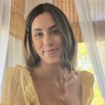 A woman with shoulder-length brown hair and a yellow top smiles softly indoors, with sunlight streaming through large windows covered by bamboo blinds and white curtains in the background.