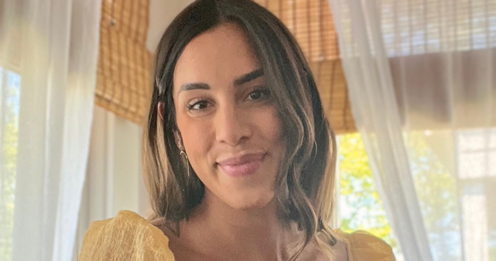 A woman with long, dark hair and a light yellow top smiles softly at the camera. She is indoors, with sheer white curtains and bamboo shades in the background. Sunlight filters through the window.