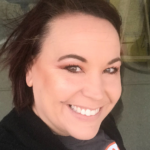 A woman with short brown hair smiles at the camera outdoors. She is wearing a black sweater and a dark top, with light makeup and a natural background behind her.