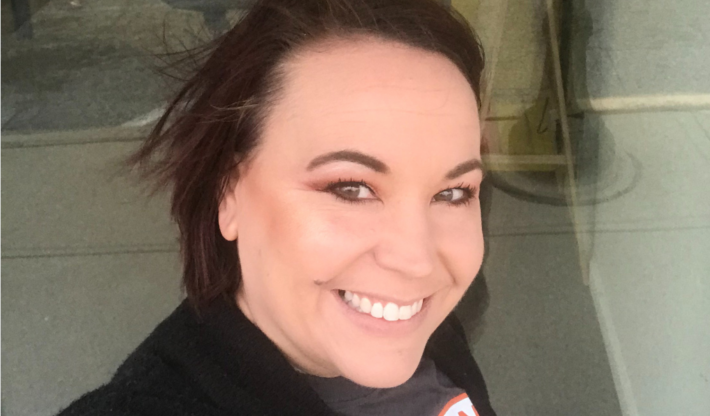 A woman with short brown hair smiles at the camera outdoors. She is wearing a black sweater and a dark top, with light makeup and a natural background behind her.