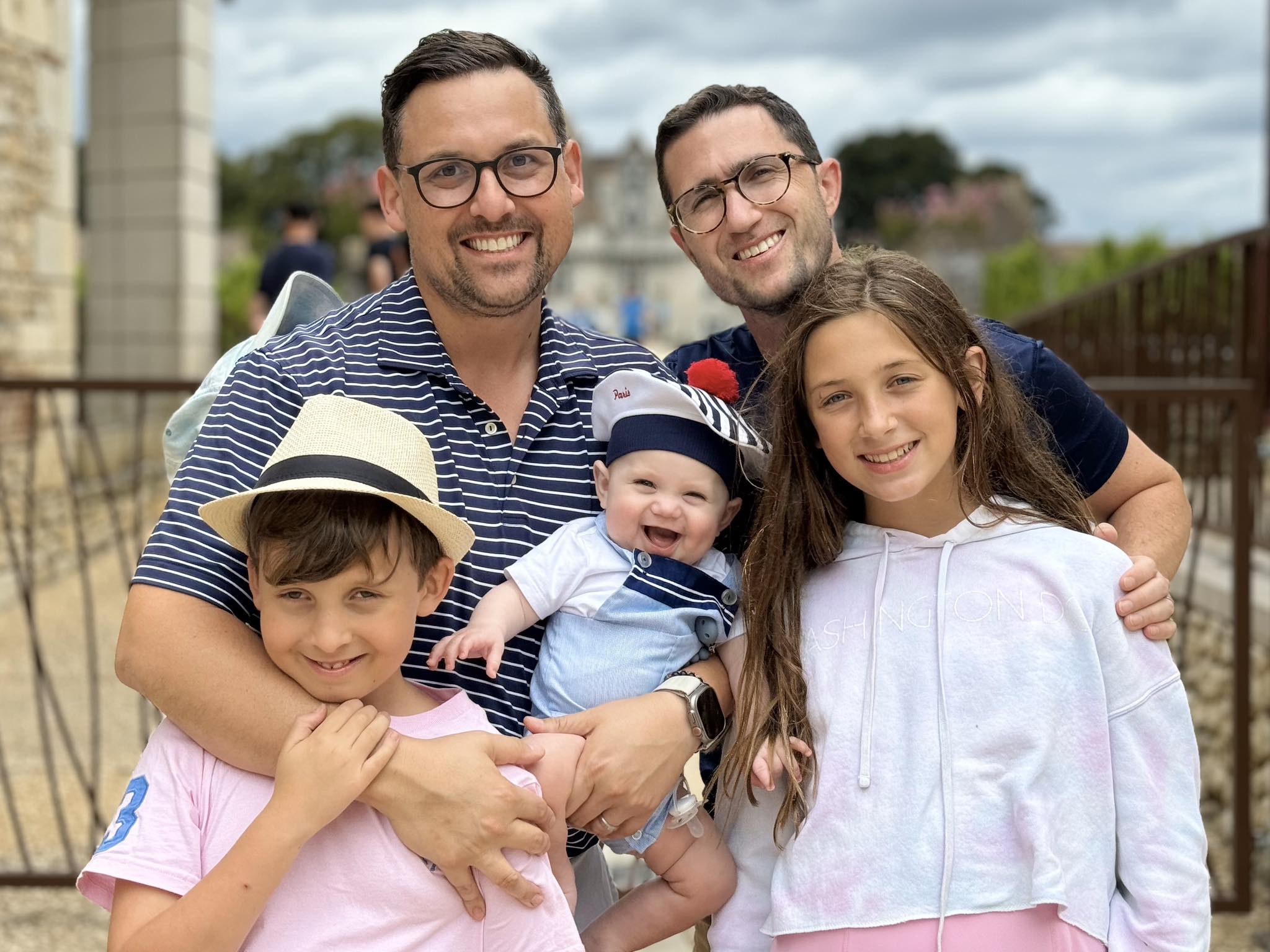 Two men and three children smile outdoors. One man holds a baby in a sailor outfit, while the other man and two older children stand close, all appearing happy. Trees and a fence are visible in the background.