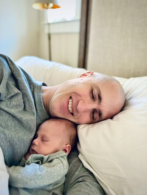 A smiling man lies in bed, cuddling a sleeping newborn baby. Both wear grey clothing and rest on white pillows, creating a peaceful and intimate scene.