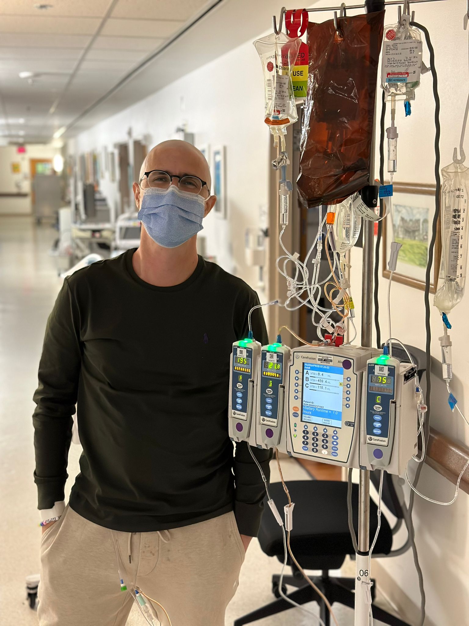 A man wearing glasses, a blue face mask, and casual clothing stands in a hospital hallway beside an IV pole with multiple infusion pumps and IV bags. He has his hands in his pockets and appears to be receiving treatment.