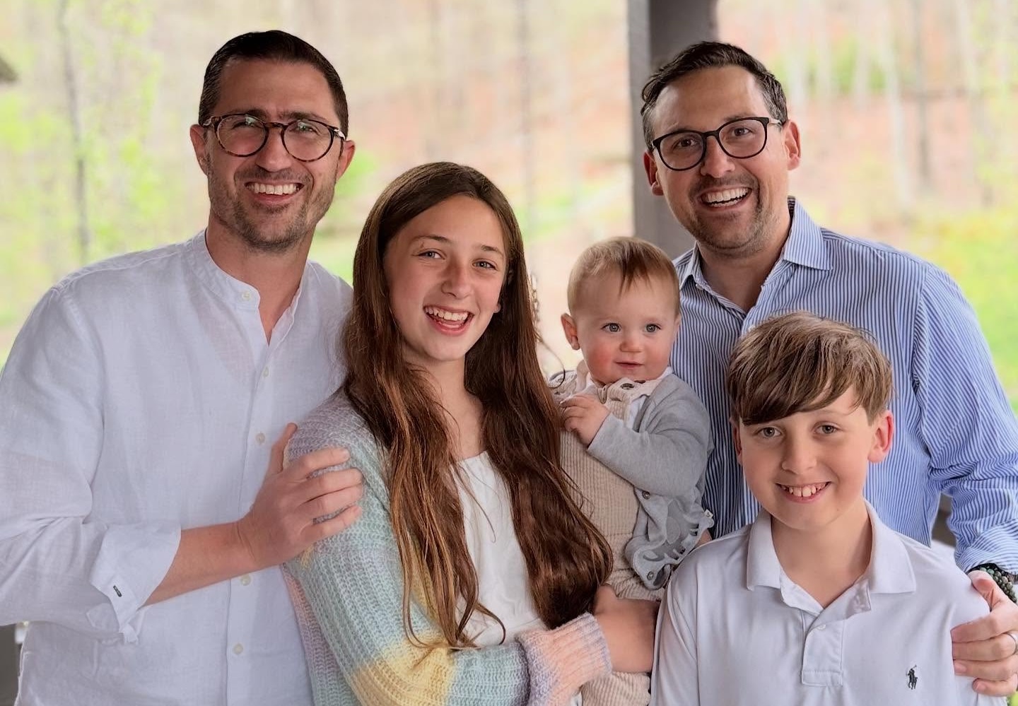 Two men stand with three children, including a teenage girl, a young boy, and a baby, all smiling and posing together outdoors in a wooded area.