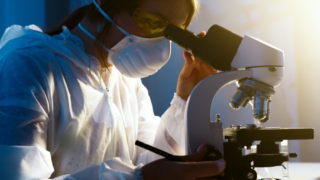 A scientist wearing protective clothing, a mask, and safety glasses looks through a microscope in a laboratory setting, with a pen in hand and focused on her research.